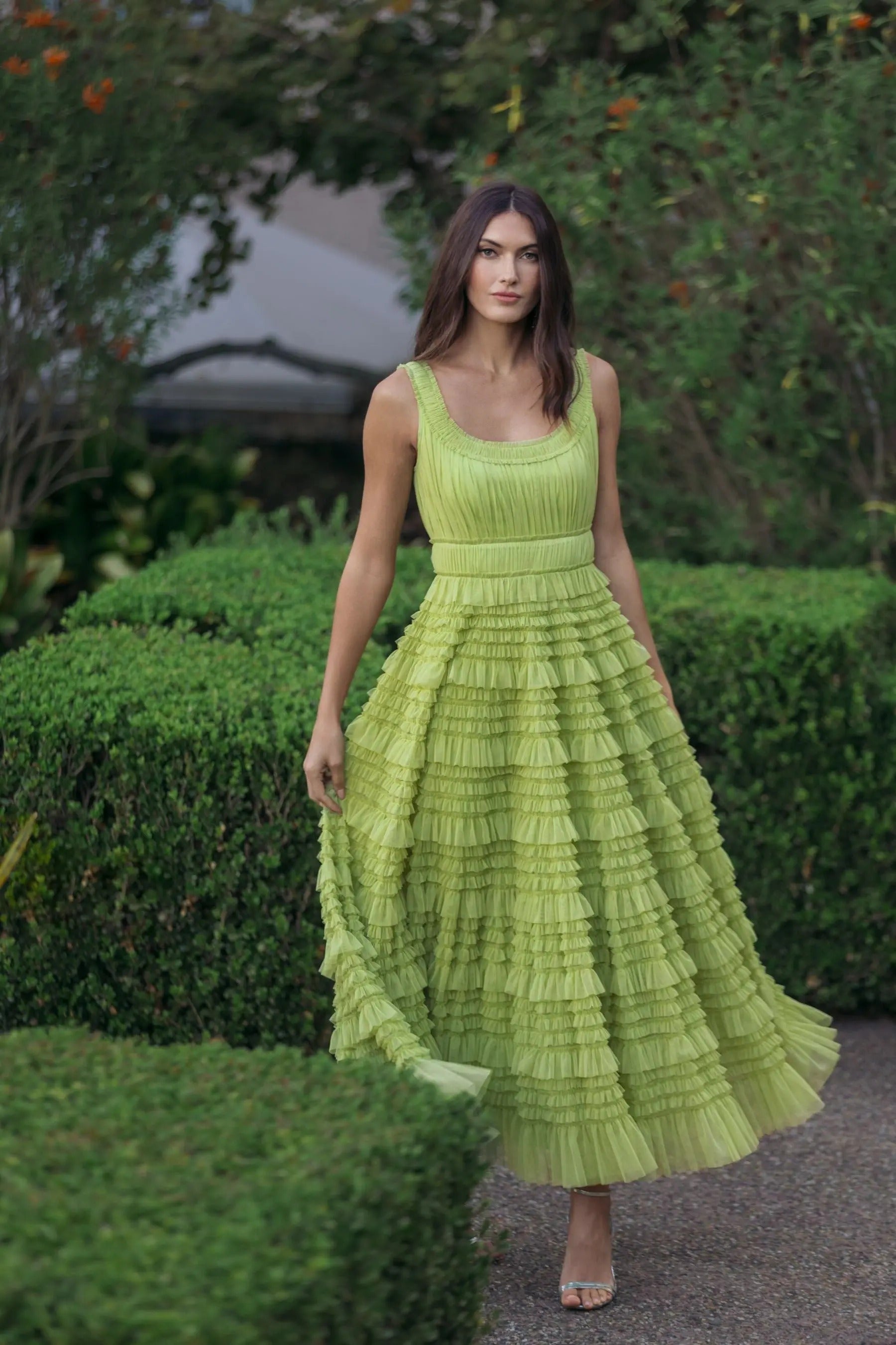 A woman wearing the Mac Duggal 8292 Tea-Length Party Dress in apple green, featuring a sleeveless, ruffled mesh design with an A-line tiered skirt, stands outdoors on a path surrounded by trimmed green bushes and trees.